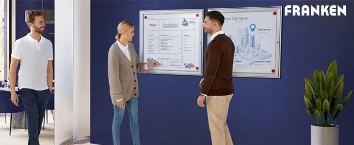Das Bild zeigt drei Personen in einem modernen, hellen Raum, die sich an einer Wand mit Plakatrahmen informieren. Die Beschilderung in den Rahmen präsentiert klar strukturierte Informationen. Ein Infodisplay im Vordergrund dient als Wegweiser und ergänzt die Orientierungshilfe. 