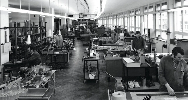 Historische Produktionshalle mit mehreren Mitarbeitenden an Arbeitsplätzen und Fertigungsmaschinen.