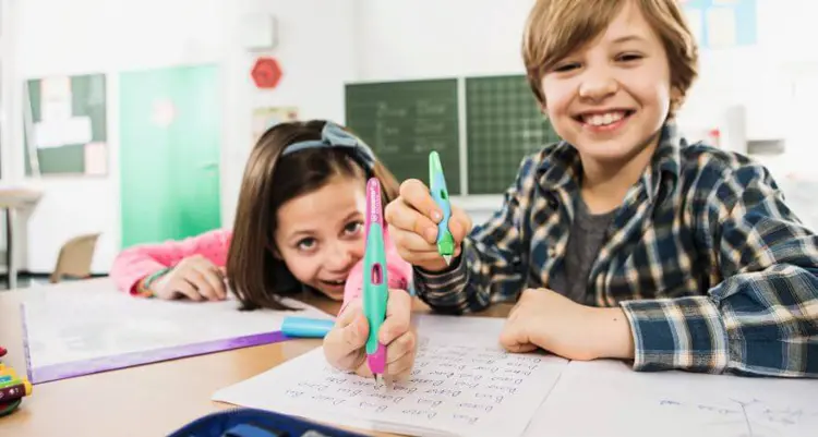 Zwei Kinder sitzen an einer Schulbank und halten lächelnd Schreiblernfüller für Rechts- und Linkshänder Richtung Kamera. 