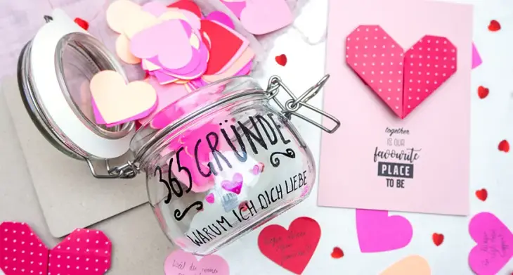 Auf einem hellen Untergrund liegt ein geöffnetes Vorratsglas, auf das mit einem schwarzen Glass & Porcelain Pen von Knorr Prandell "365 Gründe, warum ich dich liebe" geschrieben wurde. Aus dem Glas fallen viele Papierherzen in Rosa- und Rottönen. Rechts daneben liegt eine Valentinstagskarte auf die eine rotes Origami-Herz aus Papier geklebt wurde. Darunter ein Schriftzug: "together is our favourite place to be". Foto: Johanna Rundel 