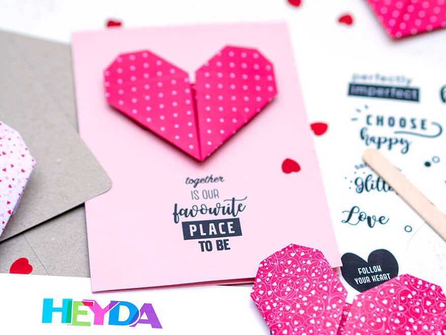 Auf einem hellen Untergrund liegt eine Valentinstagskarte, auf die eine rotes Origami-Herz aus Papier geklebt wurde. Darunter ein Schriftzug: "together is our favourite place to be" der mit den Rubbelstickern von Heyda aufgebracht wurde. Foto: Johanna Rundel