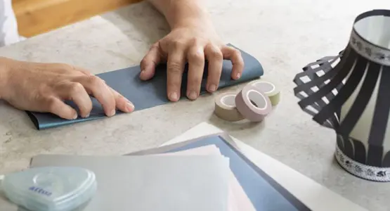 Hände falten blaues Papier. Am Rand ist eine selbstgebastelte Laterne, Bastelpapiere und Maskingtape zu sehen.