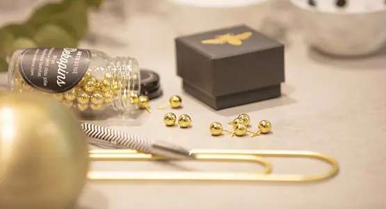 Detailaufnahme goldener Dekopins, einer goldener Büroklammer und einer schwarz-goldenen Schachtel. 