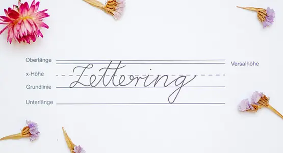 Auf einem Handlettering-Übungsblatt sind Orientierungslinien gezogen, darauf steht in Schreibschrift das Wort "Lettering", drumherum liegen Blüten.