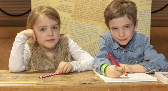 Zwei Kinder sitzen an einer Schulbank und blicken in die Kamera