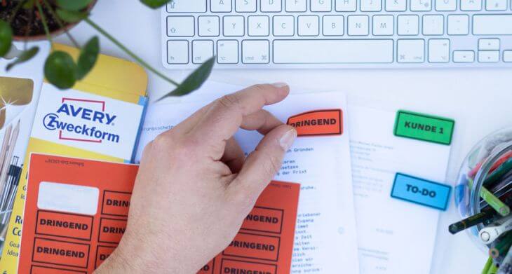 Ein Schreibtisch ist in der Draufsicht zu sehen. Eine Hand beklebt verschiedene Klarsichtfolien mit Etiketten von Avery Zweckform. Diese sind beschriftet mit "Dringend", "Kunde 1" und "To-do". Im oberen Bildrand sind eine Pflanze und eine Tastatur zu sehen. Unten rechts ist ein gefüllter Stiftebecher zu sehen.