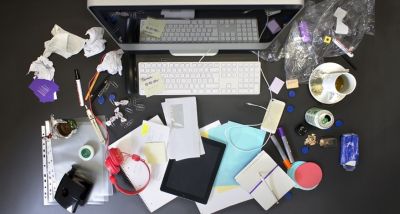 Ein unaufgeräumter Schreibtisch wurde aus der Vogelperspektive abgebildet. Man sieht verschiedene Büroutensielien wie Stifte, Papiere und Getränke um einen PC verstreut. Der Untergrund ist schwarz.