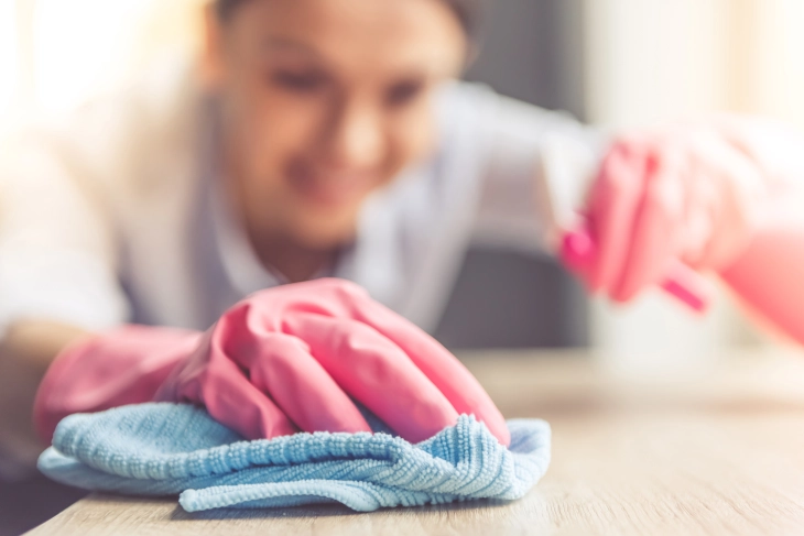 Ein Bild, das eine junge Frau in pinken Gummihandschuhen zeigt, die mit einem Tuch und einem Reinigungsspray einen Schreibtisch reinigt.