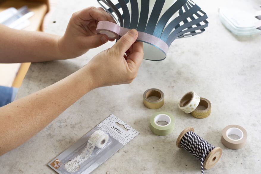 Hände umwickeln eine DIY-Laterne aus Papier mit Washitape. Auf dem Schreibtisch liegen verschiedene Bastelmaterialien von Artoz.