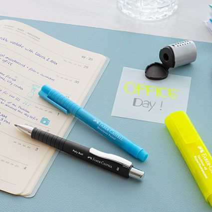 Verschiedene Stifte von Faber-Castell, ein Spitzer, Kalenderbuch und Post-it auf einer Schreibtischunterlage.