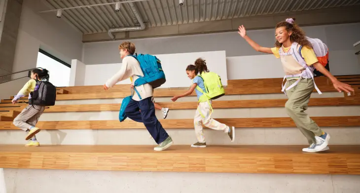 Fünf Kinder mit Schulrucksäcken von satch rennen versetzt auf Treppenstufen einer Turnhalle.
