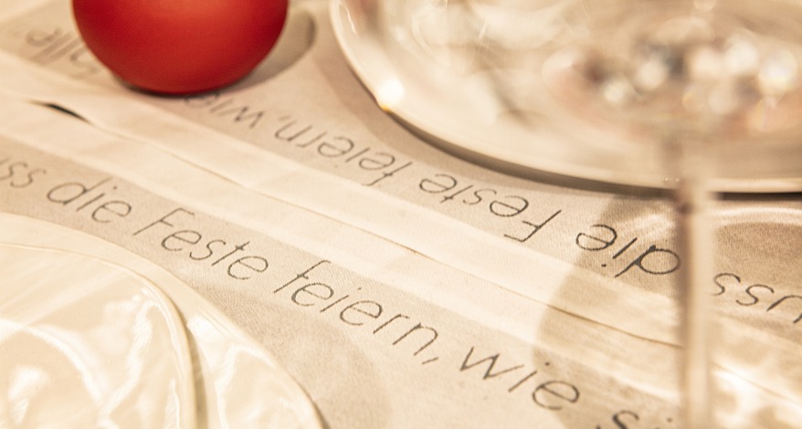 Nahaufnahme eines Tischsets mit der Aufschrift "Man muss die Feste feiern wie sie fallen". Darauf stehen Teller und ein Weinglas.