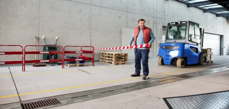 Ein Mann in einer rot-grauen Jacke hält ein rot-weiß gestreiftes tesa Absperrband in einer Werkshalle. Im Hintergrund sind ein blauer Gabelstapler und ein Stapel Holzpaletten zu sehen, außerdem ein rotes Geländer und einige Geräte.