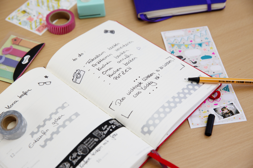In einem aufgeschlagenen Notizbuch sind die Seiten mit Terminen beschrieben und mit Maskingtape und Icons schön gestaltet. Auf dem Tisch drumherum liegen Maskingtape, Sticker, Post-its.