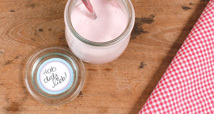 Aufnahme aus der Vogelperspektive auf einen Holztisch, auf dem ein kleines geöffnetes Weckglas steht. Auf dem Deckel klebt ein Etikett der Marke Avery Zweckform mit der Aufschrift "Hab dich lieb!". Daneben ist ein Stück eines rot-weiß karierten Tischtuchs zu sehen.