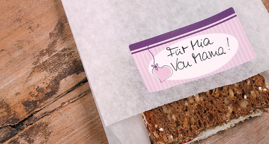 Auf einem Holztisch liegt eine mit einem Pausenbrot gefüllte Butterbrot-Tüte. Auf der Tüte ist ein lila Sticker mit Herz der Marke Avery Zweckform Living aufgeklebt. Auf dem Sticker steht geschrieben "Für Mia von Mama!"