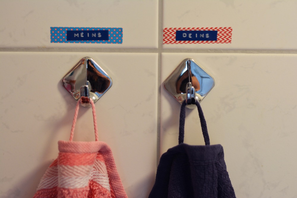 An zwei silberfarbenen Haken, die an einer Fließenwand angebracht sind, hängen zwei Hantücher. Über den Haken wurde Maskingtape und Prägeband mit der Aufschrift "Deins" und "Meins" angebracht.