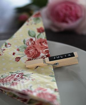 Auf einem weißen Teller liegt eine Servierte mit Blumenmuster. Diese wird von einer Holzklammer zusammengehalten. Diese wurde mit einem Prägeband mit der Prägung "Julia" beschriftet und dient somit als Platzkarte