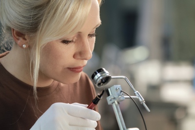 Eine Frau trägt einen weißen Handschuh und hält einen Füllfederhalter unter eine Lupe, durch die Sie schaut.