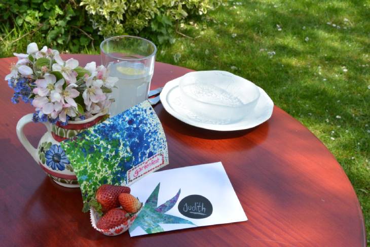 Auf einem Holztisch im Garten stehen ein Teller, ein Wasserglas gefüllt mit Limonade, eine Porzellankanne, in der Blumen stecken und es liegen 2 bunte Einladungskarten auf dem Tisch.