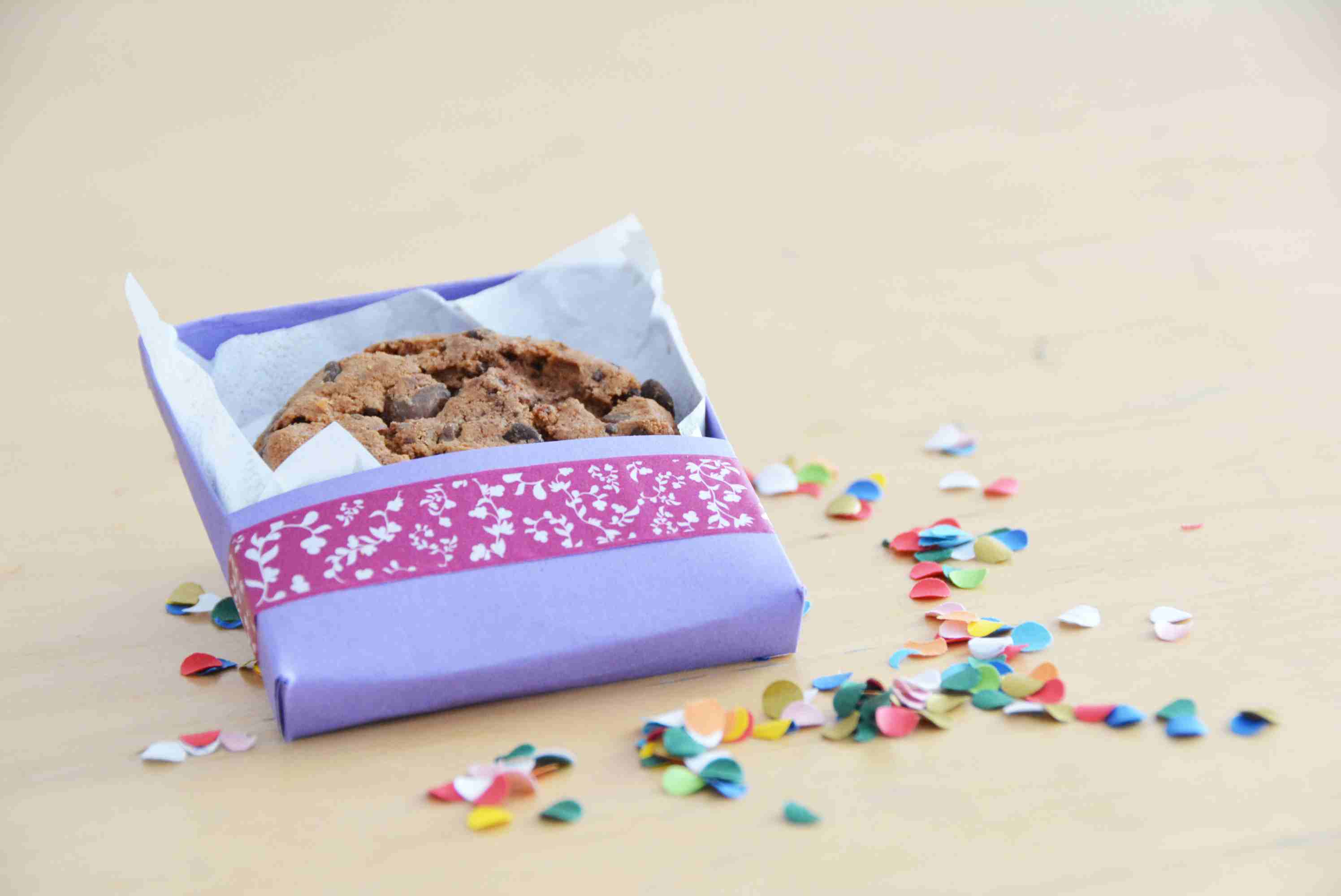 Selbstgebastelte Schachtel für einzelne Kekse. Die Schachtel wurde aus lila Karton und Washitape erstellt. In der Schachtel ist ein Keks und um die Schachtel liegt buntes Konfetti.