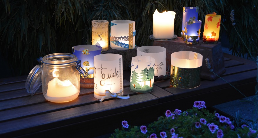 Auf einer Bank im Garten stehen im Halbdunkel viele selbstgebastelte Windlichter und leuchten.