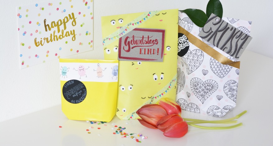 Drei selbstgebastelte Geschenktüten aus Papier stehen vor einem weißen Hintergrund. Davor liegen buntes Konfetti und rote Tulpen. Im Hintergrund ist links eine Karte mit den Worten "Happy Birthday" angebracht."