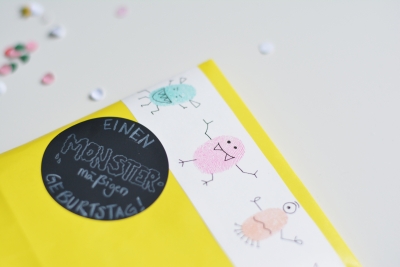 Eine gelbe DIY-Papiertüte liegt auf einem weißen Untergrund. im Hintergrund erkennt man unscharf buntes Konfetti. Die gelbe Tüte ist mit einem schwarzen Aufkleber beklebt, der mit den Worten "Einen monstermäßigen Geburtstag" belettert ist. Darüber sieht man auf einem weißen Streifen kleine, tanzende Monster, die aus Fingerabdrücken gestaltet wurden.