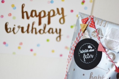 Im rechten Bildrand ist eine selbst gebastelte Papiertüte in silber zu sehen. Sie ist mit einer Wimpelkette behangen und darauf klebt ein Sitcker mit den Worten "klein aber fein". Im Hintergrund sieht man eine Postkarte mit bunten Konfetti, auf der "Happy Birthday" steht.