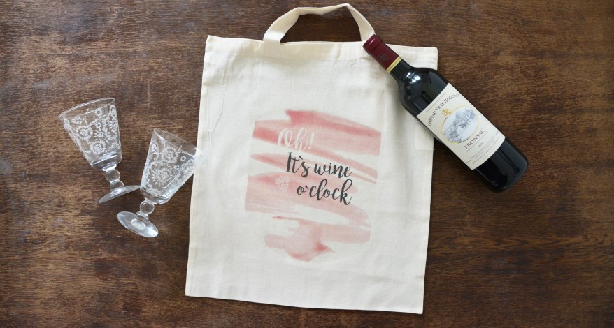 Ein bedruckter Jutebeutel liegt auf einem Tisch. Daneben liegen 2 Weingläser und eine Weinflasche.