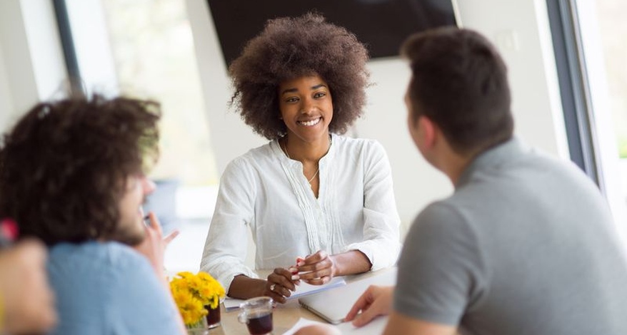 Drei Personen im Gespräch an einem Tisch, eine Person lacht freundlich.