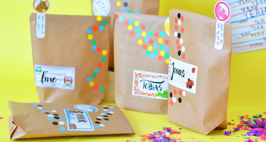 Fünf selbstgebastelte Tüten aus Kraftkarton stehen vor einem gelben Hintergrund. Am rechten Bildrand liegt Konfetti. Die Tüten wurden mit bunten Klebepunkten beklebt und mit Etiketten verziert. Die Etiketten wurden mit verschiedenen Namen belettert. 