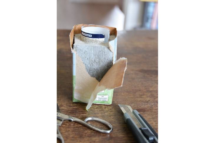 Auf einem Holztisch steht der mit Bastelbeton befüllte Milchkarton. Der Beton ist ausgehärtet und der Milchkarton wird vom Beton gelöst. Es liegt eine Schere und ein Cuttermesser auf dem Tisch.