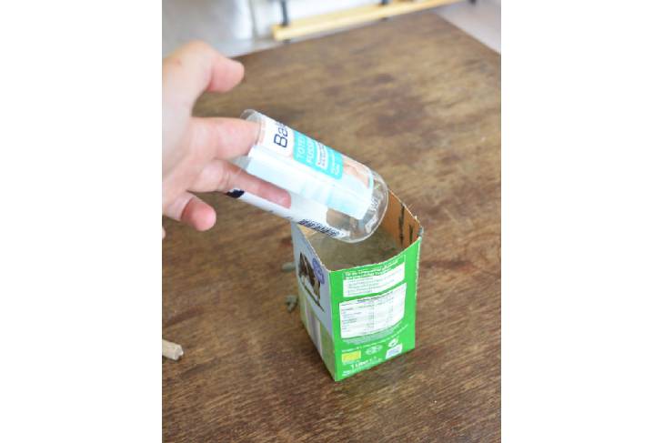 Auf einem Holztisch steht die halb mit Bastelbeton gefüllte Milchtüte. Eine Hand fügt eine Plastikflasche in den Beton ein.