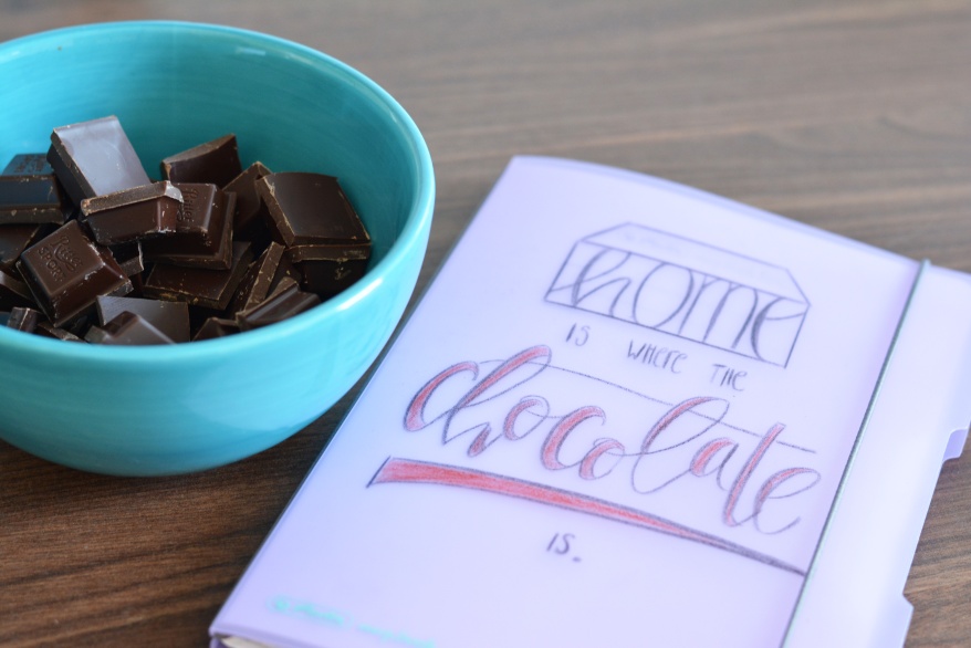 Auf einem Holztisch liegt ein Notizheft der my.book flex Reihe von Herlitz. Das Buch wurde mit den Worten "Home is where the chocolate is" belettert. Links neben dem Buch steht eine türkisfarbene Schale mit Schokoladentücken.