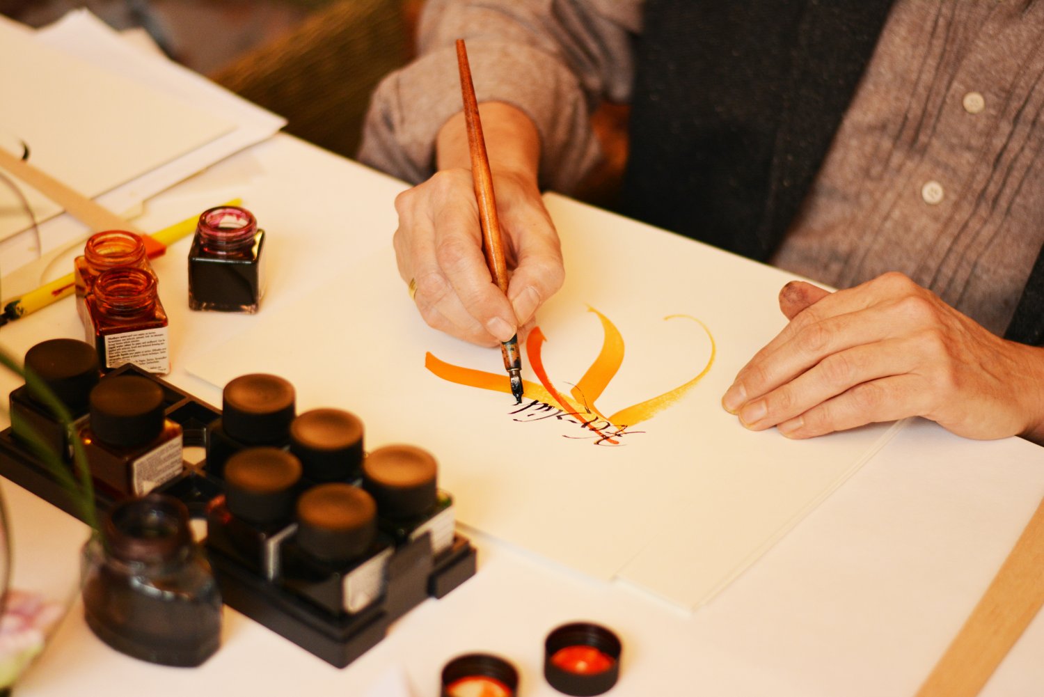 Auf einem Blatt ist ein Elementmit oranger Tinte gemalt. Darüber wird gerade etwas kalligrafiert. Es stehen viele Tintenfäaaser mit bunter Tinte auf dem Tisch.