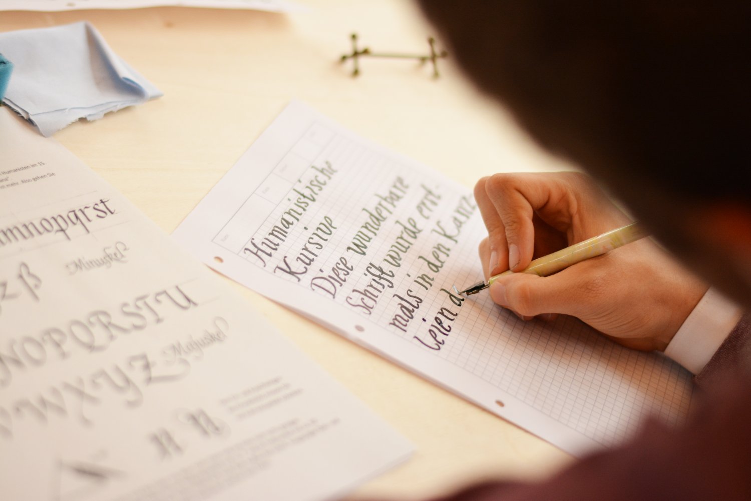 Auf einem Papier wird die Kalligrafie-Schrift "Humanistische Kursive" geübt. Daneben liegt eien Vorlage der Schrift zur Orientierung. 