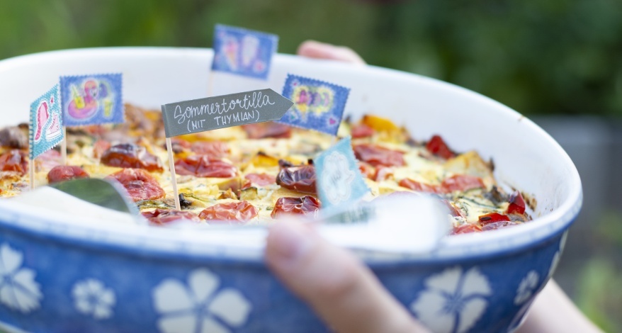Zwei Hände halten eine blaue Auflaufform mit weißen Blumen darauf. Aus dem Tortilla, der sich in der Form befindet, ragen verschiedene Fähnchen aus Washitape heraus.Ein Fähnchen ist schwarz und wurde mit weißem Stift mit den Worten "Sommertortilla mit Thymian" beschriftet.