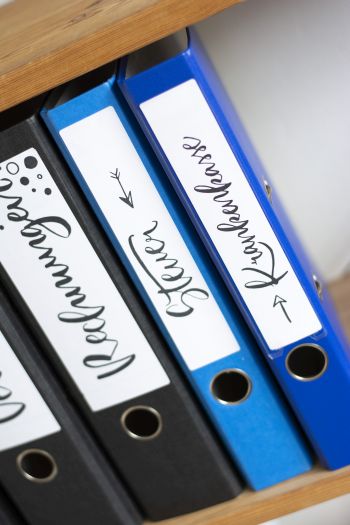 In einem Holzregal stehen mehrere Ordner, die mit Ordnerrückenetiketten der Marke Avery Zweckform beschriftet wurden. Darauf zu lesen ist "Versicherungen", "Steuer" und "Krankenkasse"