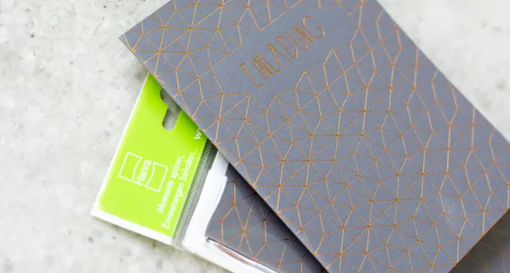 Eine graue Einladungskarte mit goldenem Muster der Marke Hanra liegt auf einem Betontisch.