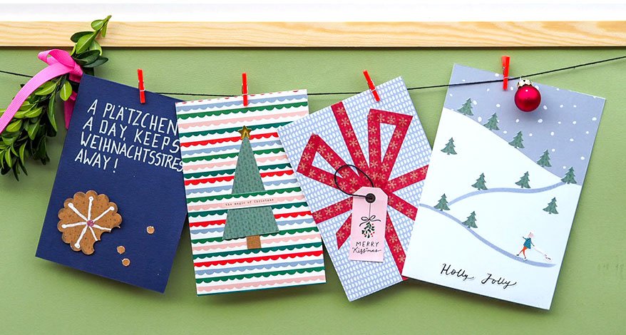 Vier selbstgebastelte Weihnachtskarten hängen an einer Schnur. Sie sind mit roten Klammern befestigt. An der Schnur sind ebenfalls eine Weihnachtskugel und ein Mistelzweig mit rosa Schleife angebracht. Der Hintergrund ist grün. 