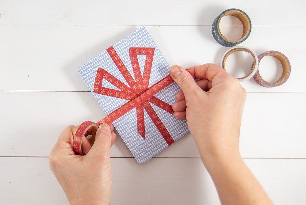Ein Motivkarton in grau-weiß wird mit rotem Washitape beklebt. Die Anordnung des Tapes ist dabei so gewählt, dass dieses wie die Schnüre um eine Geschenk aussehen. Auf der rechten Seite liegen weitere Washitape-Rollen. Der Untergrund ist weiß.