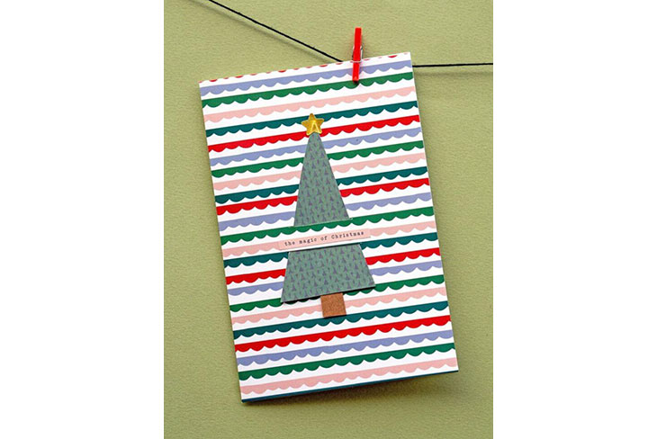 Auf einen Motivkarton mit Streifenmuster wurde ein Aufkleber mit Weihnachtsbaummotiv geklebt. Dieser wurde unter einem Streifen geschoben, auf dem "The Magic of Christmas" geschrieben wurde. Der Untergrund ist grün. Die Karte ist mit einer roten Klammer an einer Kordel befestigt."