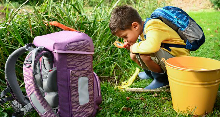 Ein Kind mit einem Schulranzen von Step by Step hockt mit einer Lupe in der Hand auf einer Wiese und beobachtet etwas