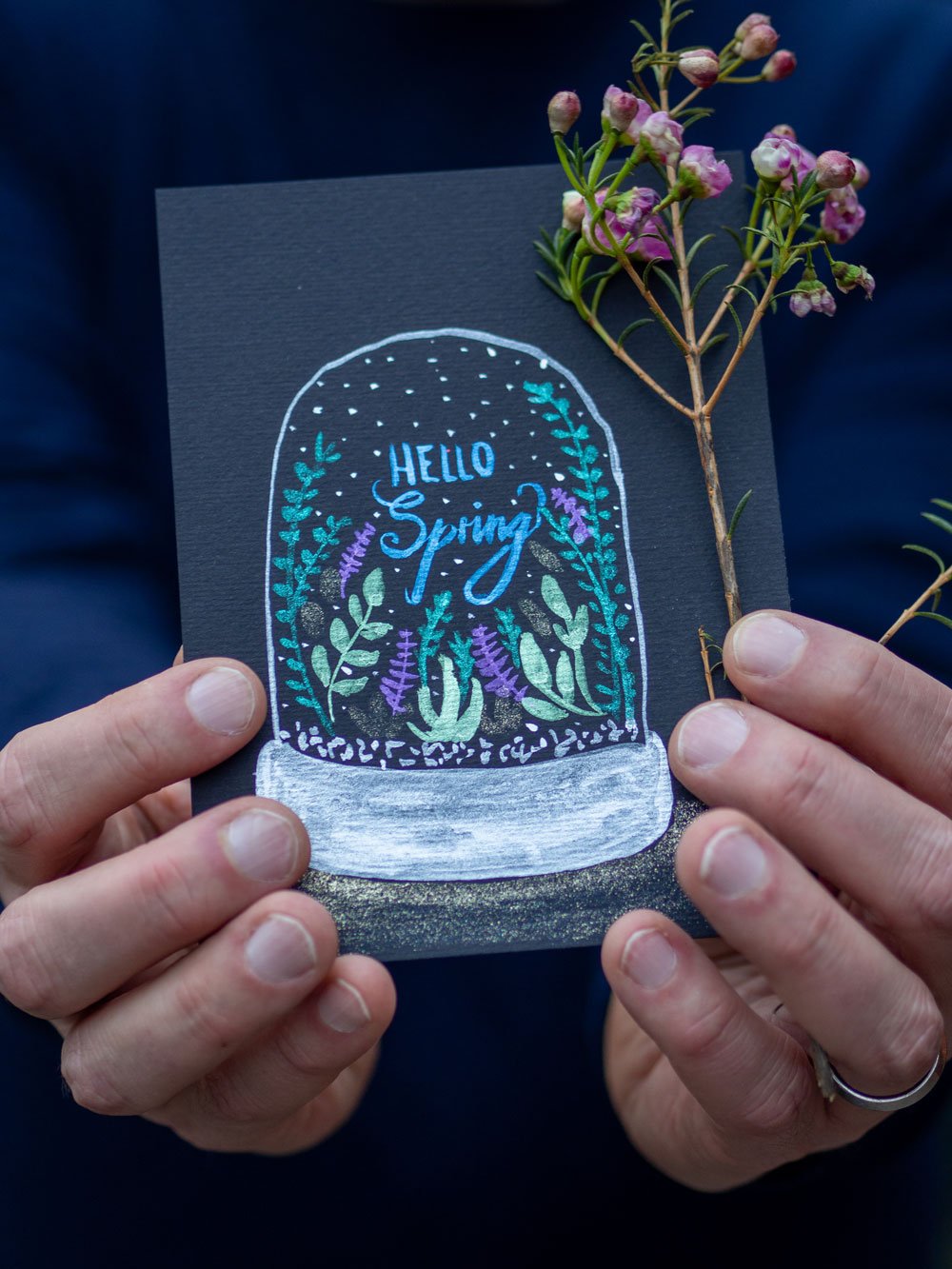 Hand hält schwarze Karte mit "Hello Spring"-Motiv und glitzernden Farben.