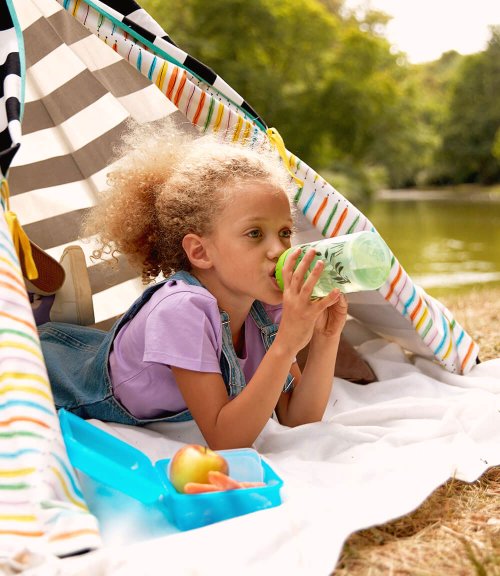 Ein Mädchen liegt auf einer Wiese in einem Zelt. Sie trinkt gerade an einer Trinkflasche, neben ihr liegt eine offene Brotdose mit Obst.