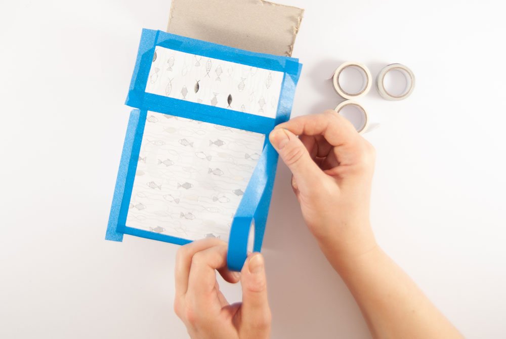 Eine Papschachtel wurde mit Motivkarton mit Fischen darauf beklebt. Zwei Hände belekleben die Kanten der Verpackung mit blauem Klebeband. Im Hintergrund sind drei Washitaperollen zu sehen.