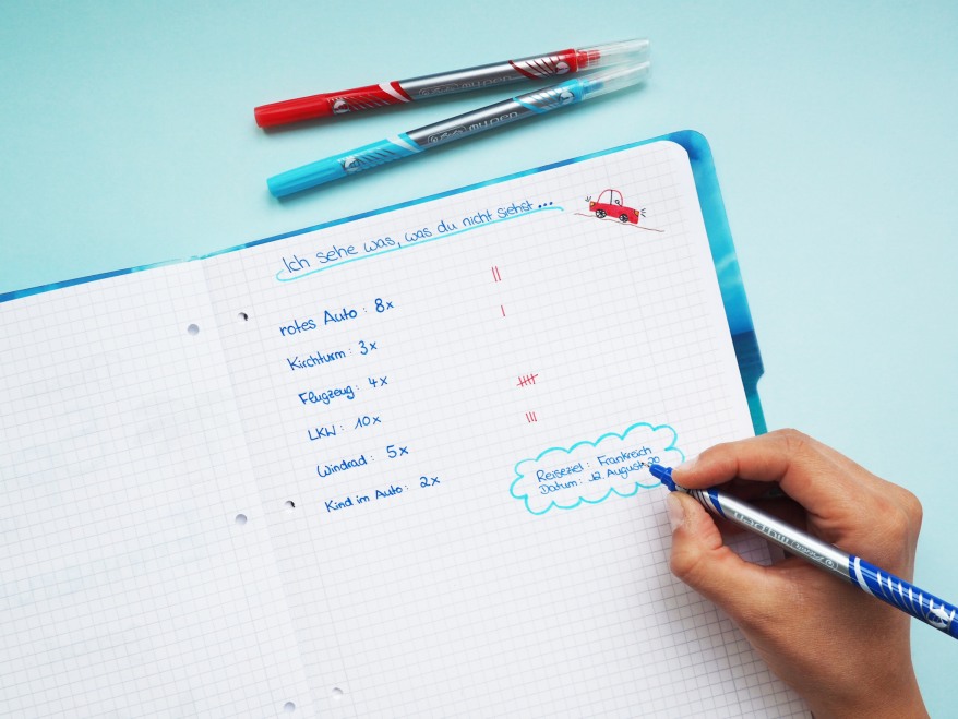 Hand schreibt Reisespiel „Ich sehe was, was du nicht siehst“ in ein Notizheft.
