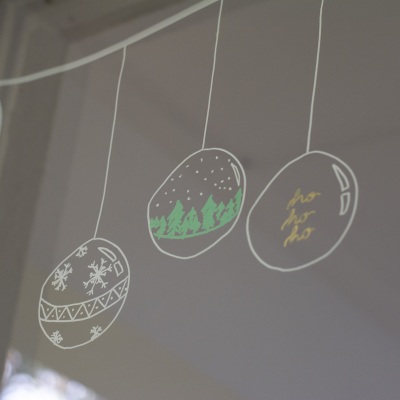 Mit weißem Kreidemarker auf ein Fenster gemalte Weihnachtskugeln vor einem dunklen Hintergrund.