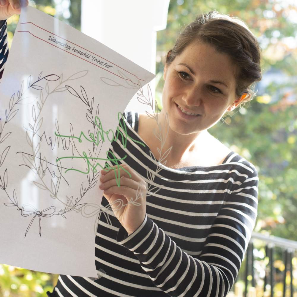 Eine junge Frau nimmt die Malvorlage für das weihnachtliche Fensterbild vom Fenster und betrachtet die Zeichnung auf dem Fenster.